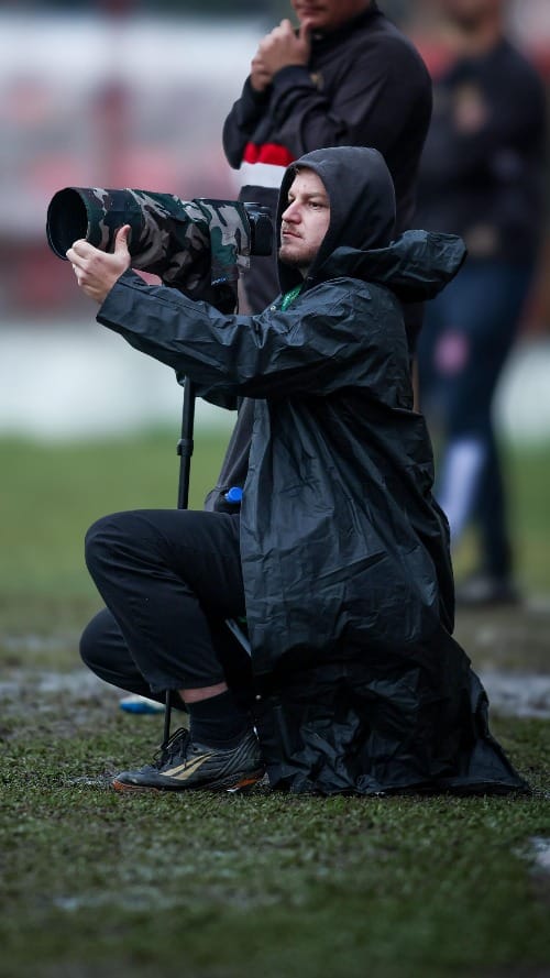 Fotógrafo Gabriel Haesbaert agachado na beira do campo, usando capa de chuva e segurando câmera.