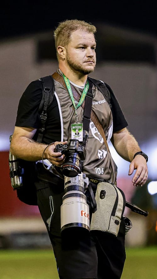 Gabriel Haesbaert caminha pelo gramado carregando câmeras e equipamentos durante cobertura fotográfica esportiva.