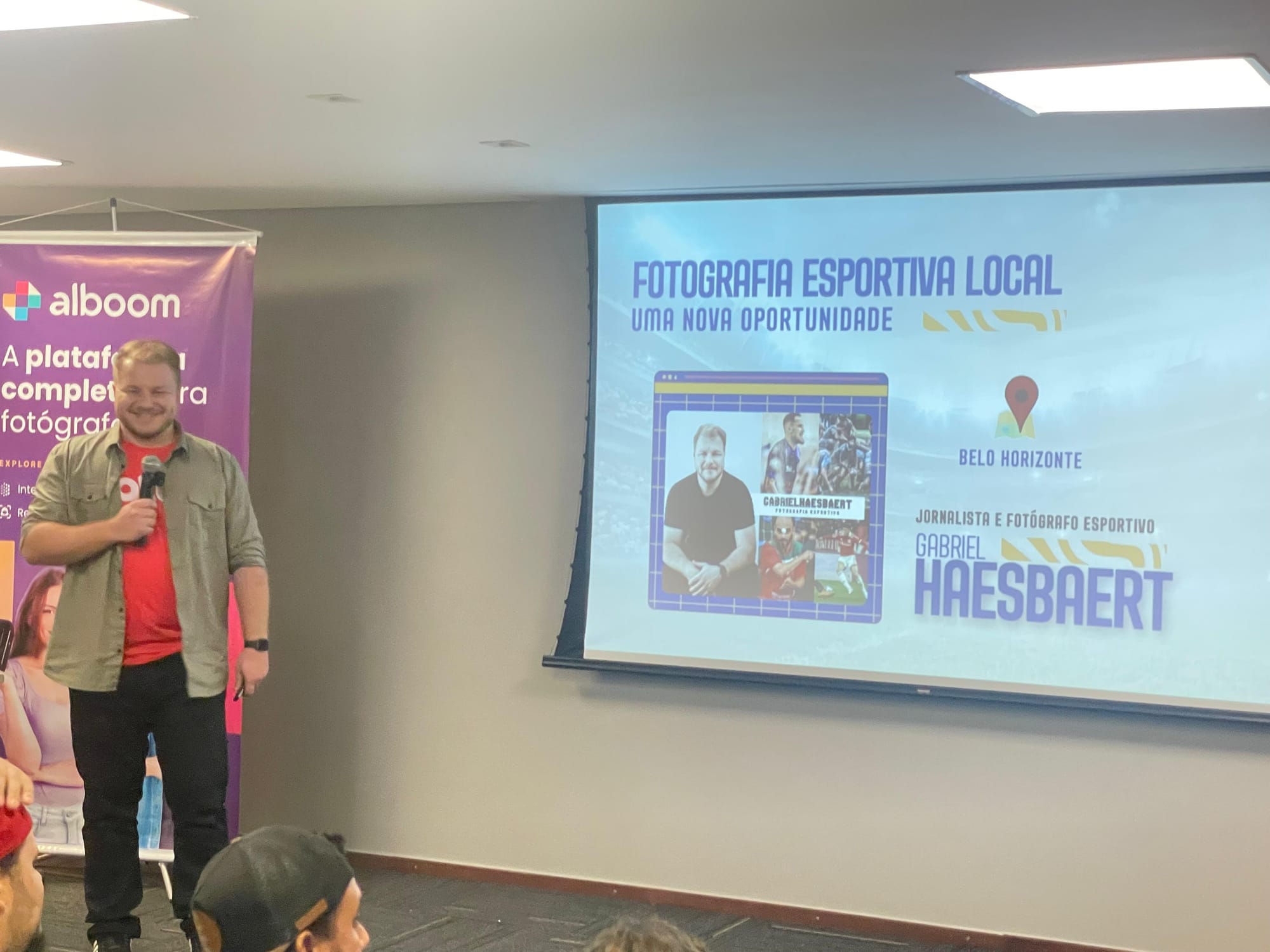 Gabriel Haesbaert sorri enquanto apresenta uma palestra sobre fotografia esportiva local em Belo Horizonte. Ele segura um microfone diante de uma tela de projeção com seu nome e imagens de seu trabalho, ao lado de um banner da plataforma Alboom.