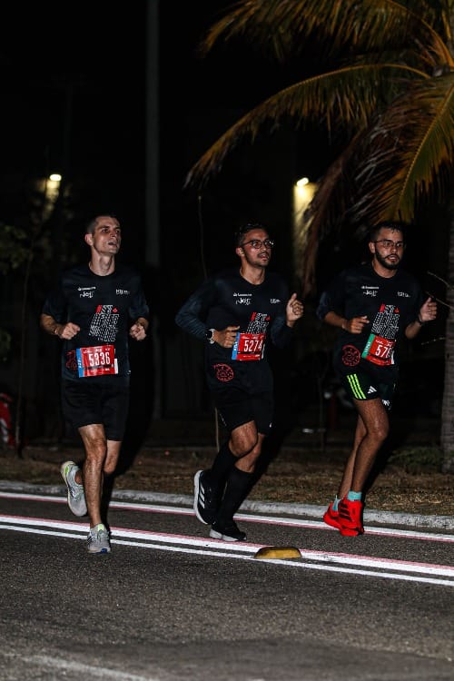 Três homens correndo à noite na corrida Night Run, com roupa esportiva preta com número de peito.