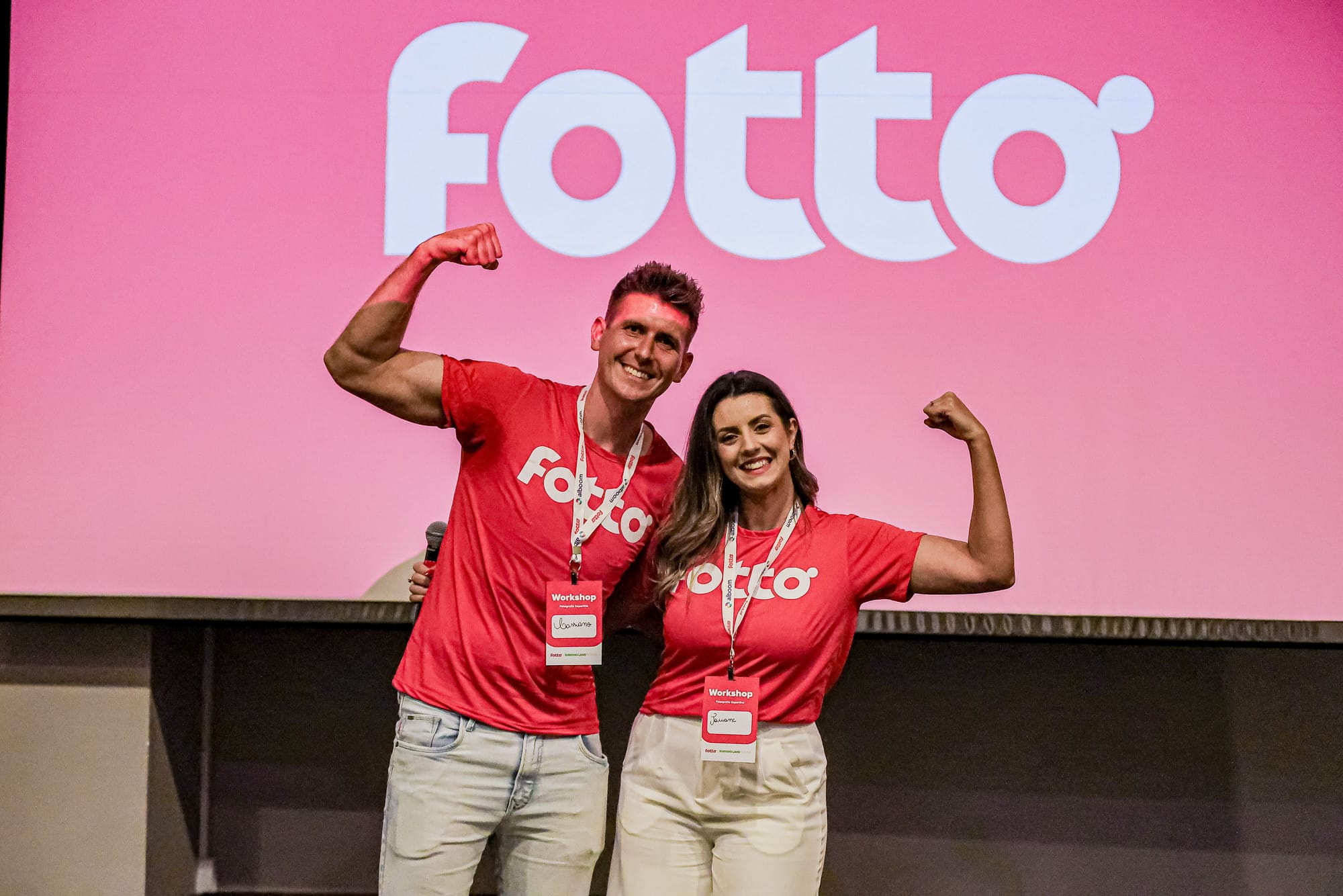 Dupla de fotógrafos posa sorridente e confiante com os braços flexionados em pose de força diante de um telão com o logo da Fotto, durante um workshop. Ambos vestem camisetas vermelhas com a logo da Fotto e crachás de identificação