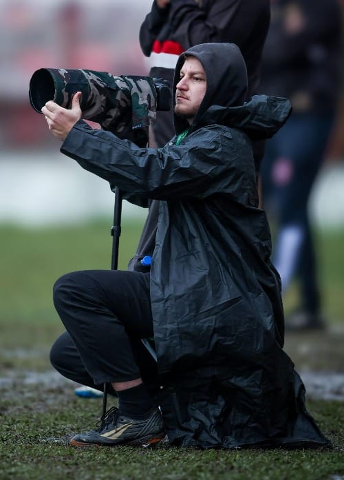Fotógrafo agachado na beira do campo, usando capa de chuva e segurando câmera fotográfica.