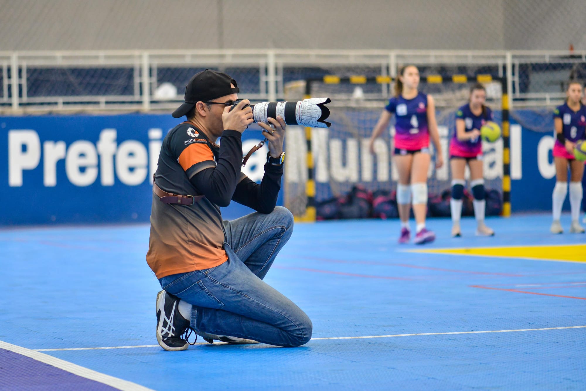 Fotógrafo ajoelhado na quadra de esportes faz registros com uma câmera, enquanto atletas de vôlei se preparam ao fundo para o jogo.