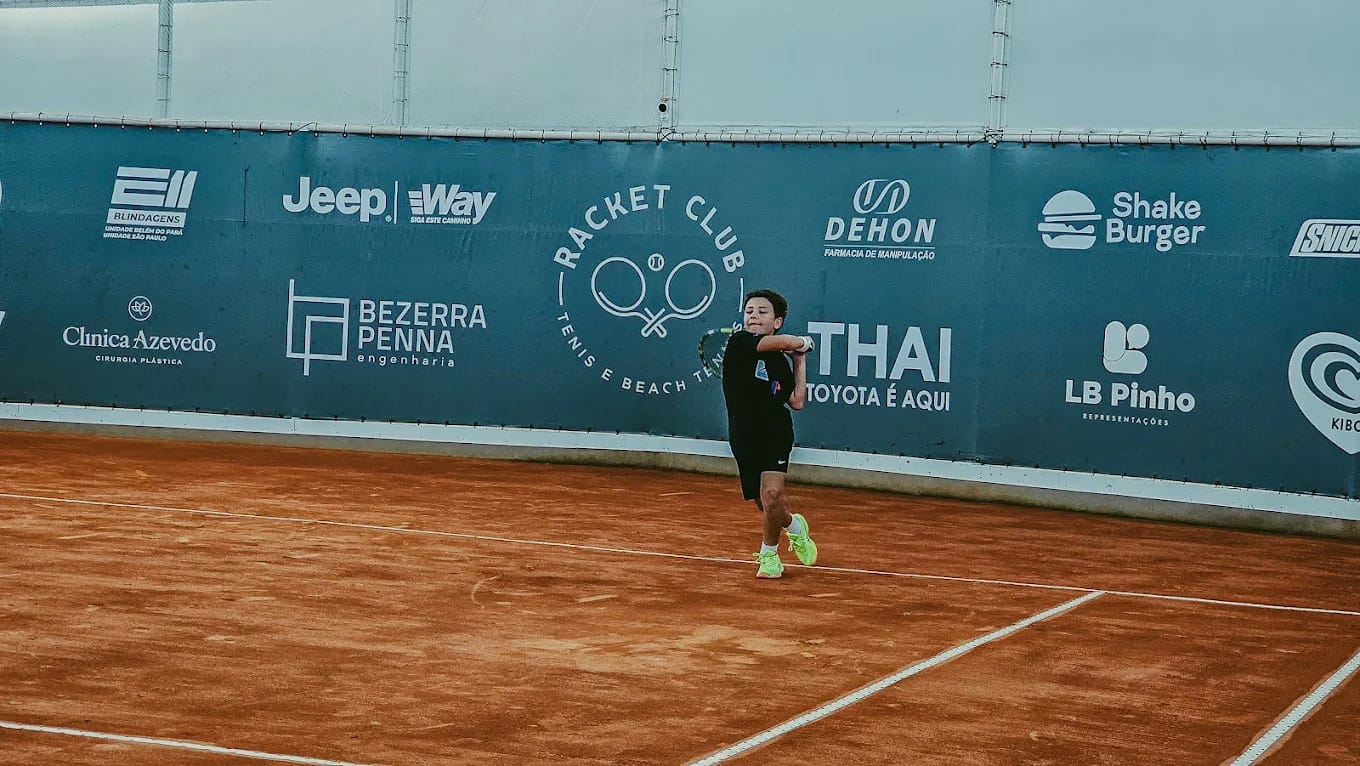 ovem atleta realiza um golpe de forehand em uma quadra de saibro coberta, durante uma partida de touch tennis. Ao fundo, o muro da quadra exibe logomarcas de diversos patrocinadores, como Jeep, Thai e Shake Burger, com o logotipo do Racket Club ao centro. O jogador veste roupas esportivas pretas e tênis verde-limão, demonstrando concentração e técnica.