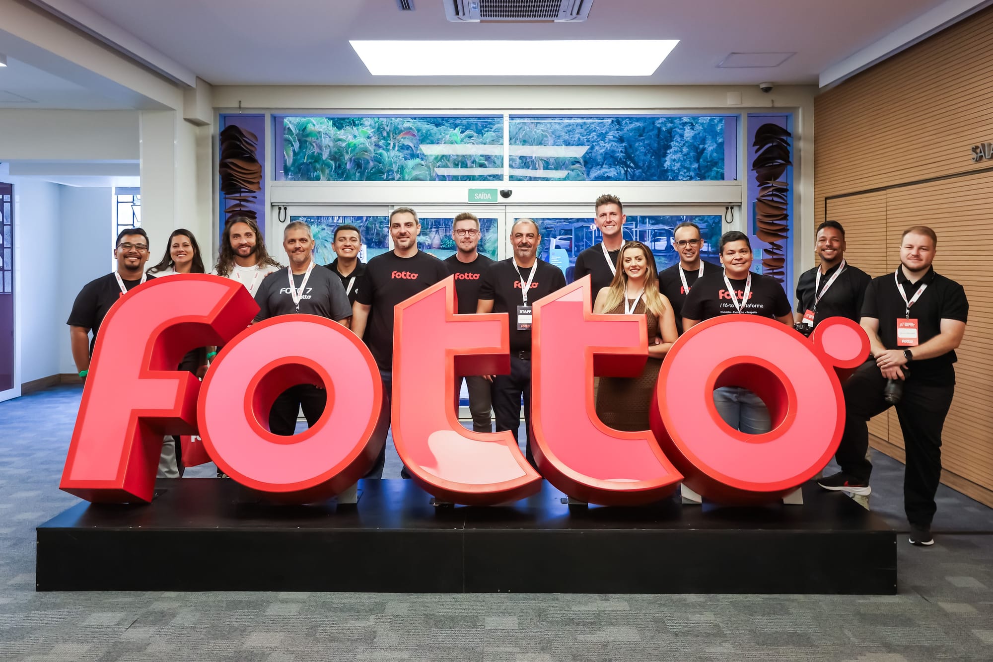 Embaixadores Fotto sorrindo em pé atrás de um letreiro grande com o nome Fotto em destaque. Todos usam crachás de identificação.