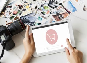 Pessoa segurando tablet com carrinho de compras visível na tela, ao lado de uma câmera profissional e várias fotos impressas.