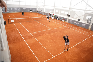 Vista superior de uma quadra de saibro indoor durante uma partida de duplas de touch tennis. Quatro jogadores estão posicionados em lados opostos da quadra, com um deles realizando o saque.